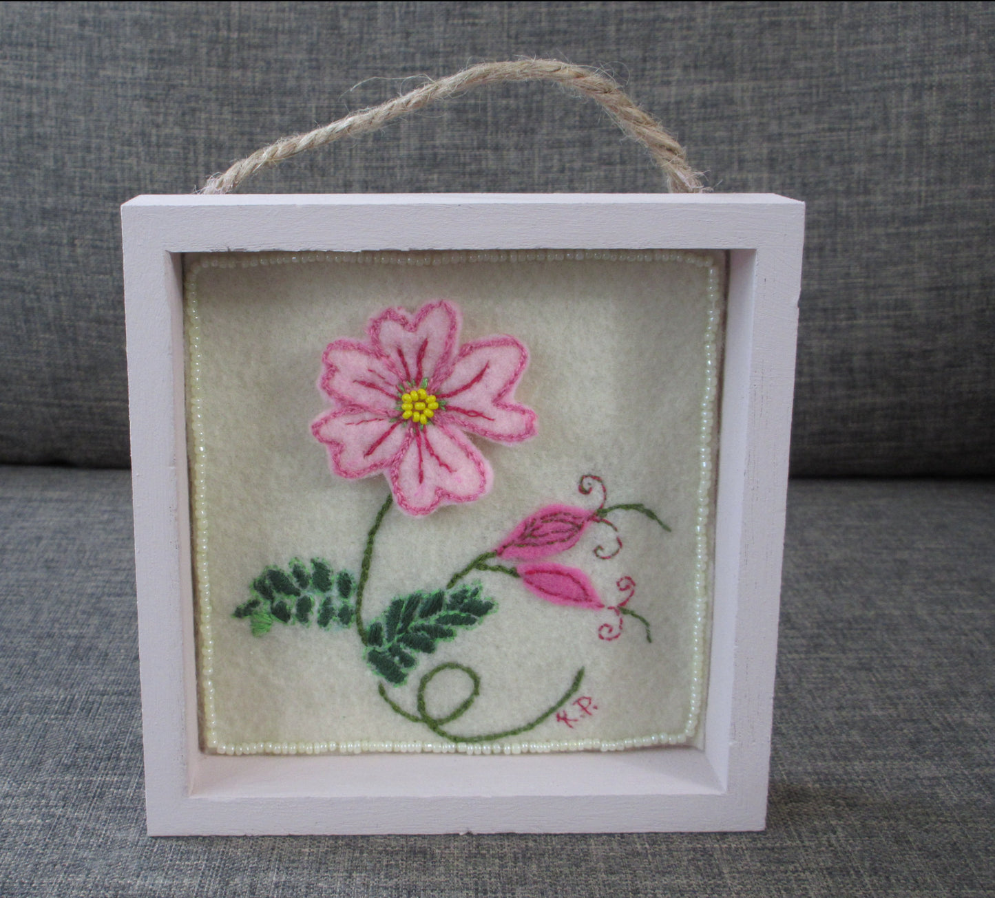 A pink flower with two buds. The flower is on felt with hand details in hand embroidery. The flower center is beads. The piece is in a chalk pink wooden box frame with a rope hanger on top. By artist Kathy Poitras.