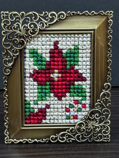 Framed cross-stitch artwork featuring a red poinsettia with green leaves , stitched with cotton and metallic threads. Folk-art style design in a small ornate gold colored frame, handmade by North Vancouver artist Kathy Poitras.
