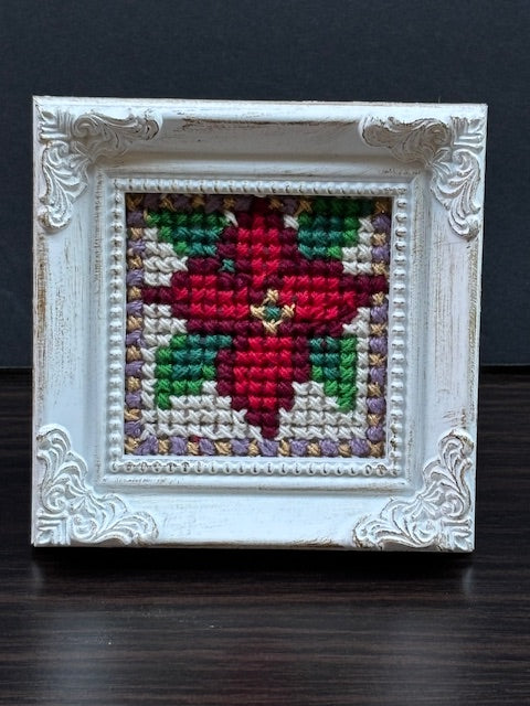 Handmade poinsettia cross-stitch in ornate white frame. Original Christmas folk-art design by North Vancouver artist Kathy Poitras.